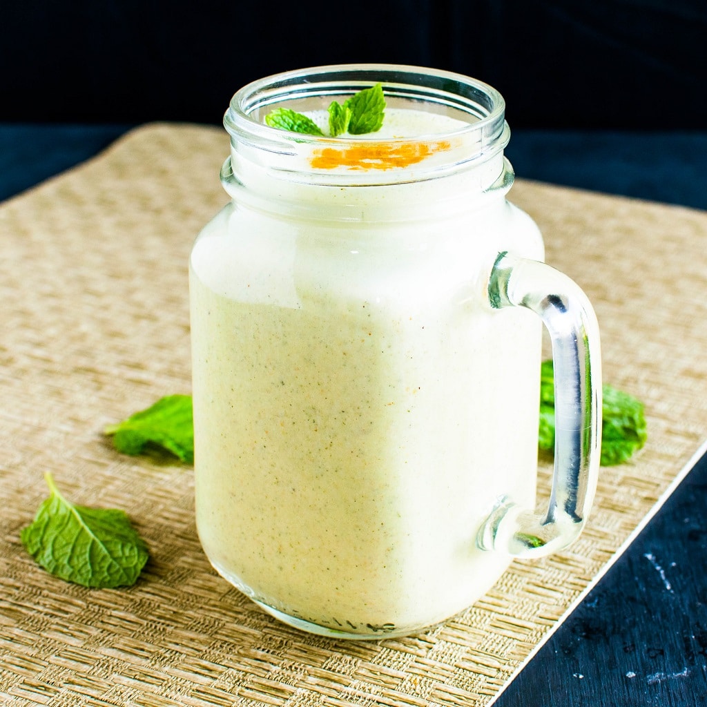 A tall glass filled with turmeric mint protein smoothie on a brown weaved mat is displayed in this image with mint leaves as the prop | kiipfit.com