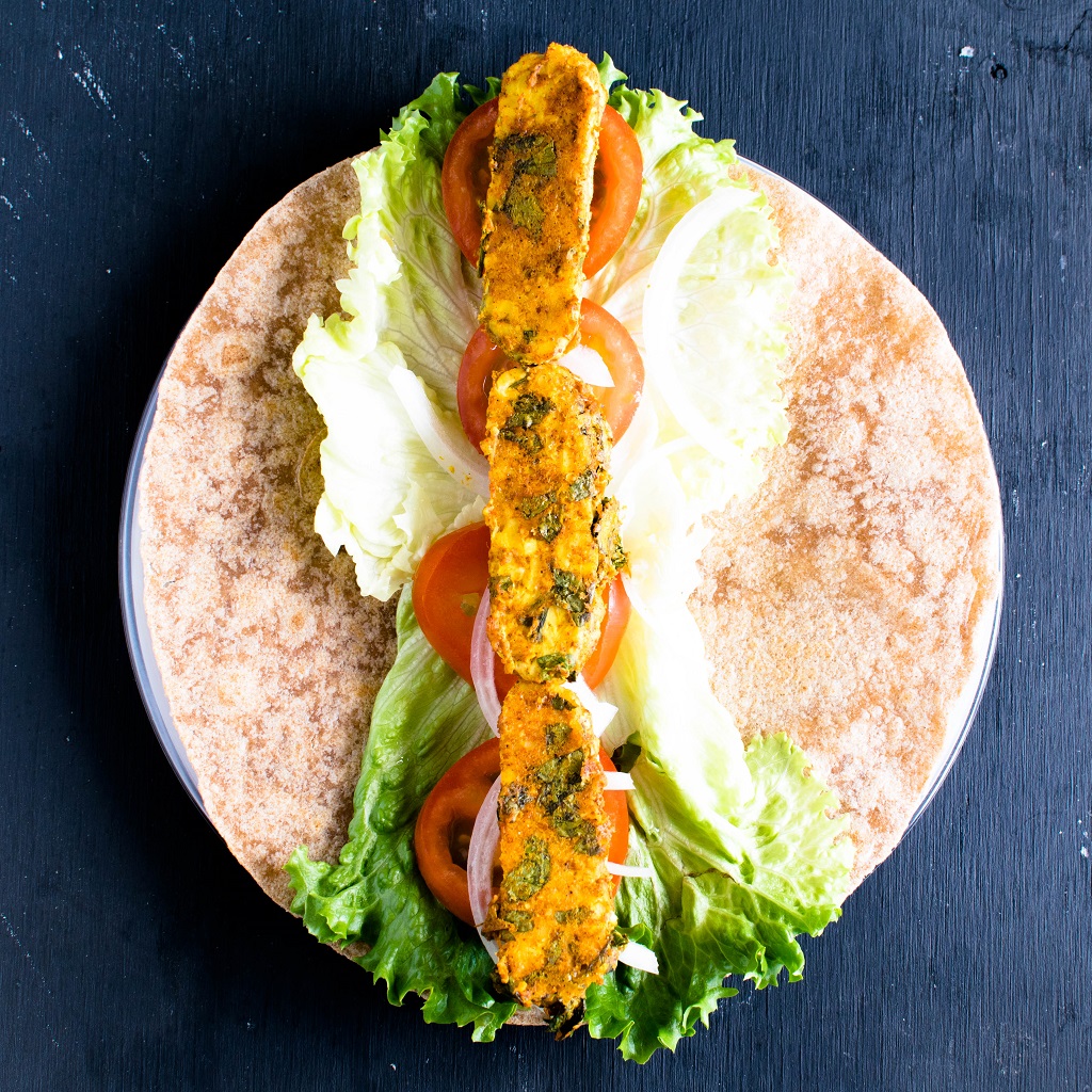 A whole grain tortilla with salad and cooked tempeh being in the process of forming a wrap is shown in this image.